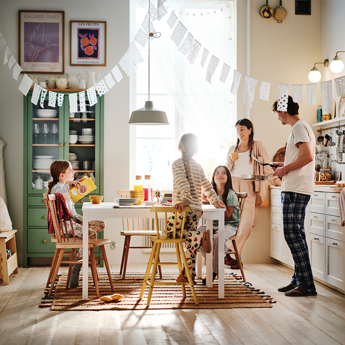 Familia desayunando en una cocina luminosa: una mesa de comedor blanca rodeada de sillas de madera natural y una trona amarilla, frente a un armario alto verde oscuro.