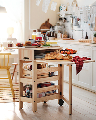 Carrito de cocina de madera de pino sin tratar con ruedas, estantes abiertos y tabla superior extensible, cargado con panes, platos y zumos.