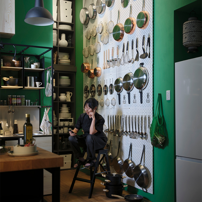 Cocina con paredes verdes: una mujer sentada en un taburete plegable negro frente a un gran panel perforado de almacenaje, con sartenes, ollas y utensilios metálicos colgados.