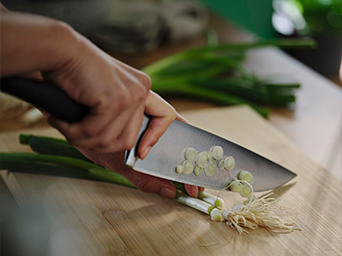 Manos usando un cuchillo de cocinero de hoja de acero inoxidable y mango negro para picar finamente cebolletas frescas sobre una tabla de cortar de bambú.