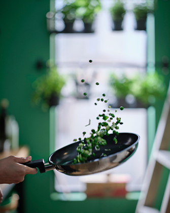 Primer plano de una mano lanzando guisantes y verduras picadas al aire con una sartén de acero con revestimiento antiadherente sobre un fondo verde con plantas.