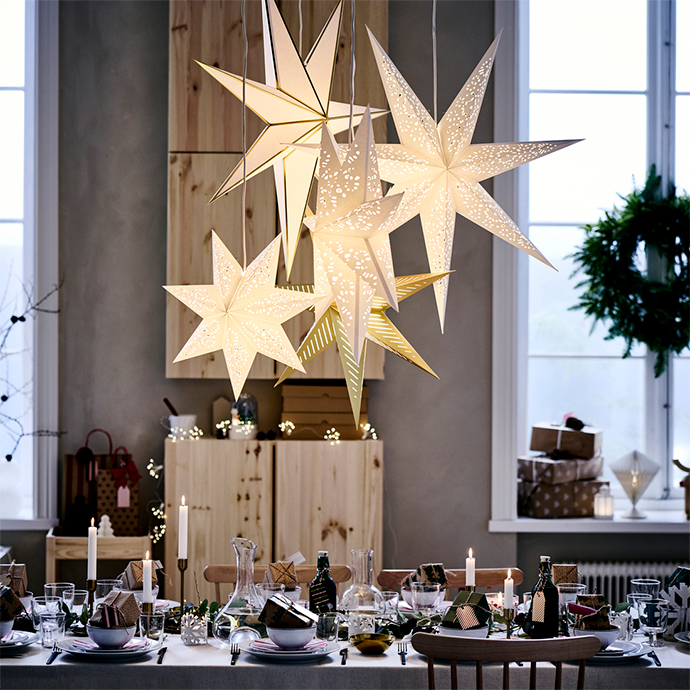 Mesa de comedor vestida para una celebración, iluminada por un grupo de cinco lámparas colgantes de papel blanco y dorado con forma de estrella perforada.