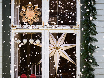 Ventana de madera blanca y desgastada, decorada con estrellas luminosas de papel colgantes y una guirnalda de abeto, con nieve cayendo en el exterior.