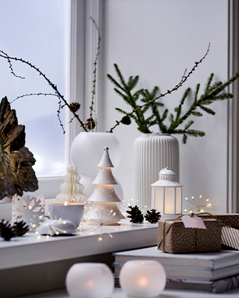 Alféizar de ventana blanco decorado con jarrones, ramas, una linterna blanca y figuras luminosas LED de cerámica blanca con forma de árbol de Navidad.