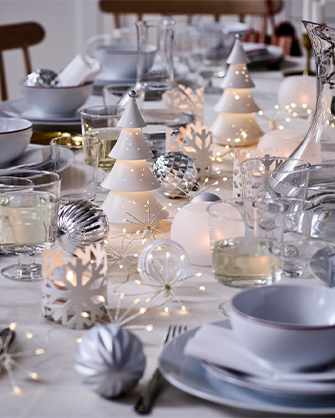 Detalle de una mesa festiva con tres figuras LED de cerámica blanca en forma de árbol y luces de hadas entre vajilla de gres gris claro y copas de cristal.
