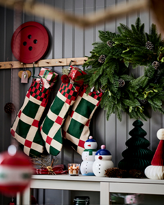 Gray plank wall decorated with three fabric Christmas stockings in red, green, and beige checker pattern, hung from a shelf above paper and ceramic decoration figures.