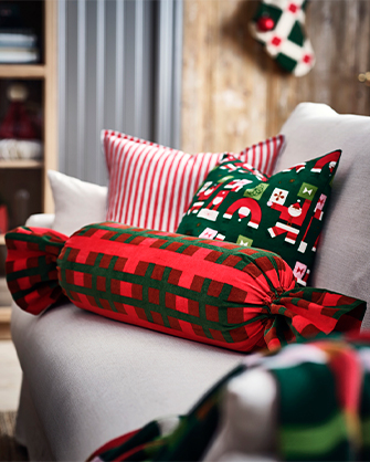 White upholstered fabric armchair with a stack of three Christmas decorative cushions in red and green, including one striped, one with a Santa pattern, and one candy-shaped bolster.