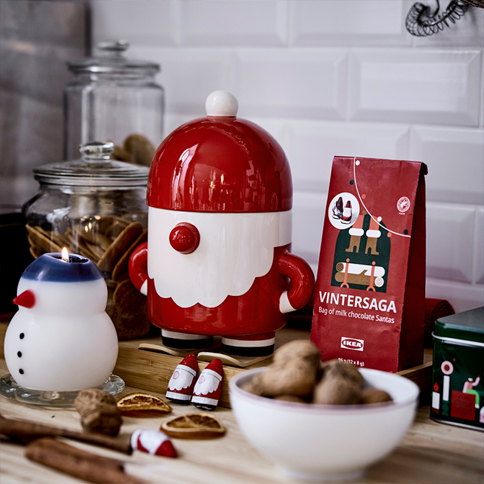 Christmas still life on a wooden countertop: a white and red stoneware container shaped like Santa Claus, a snowman candle, nuts, and a bag of VINTERSAGA series chocolates.