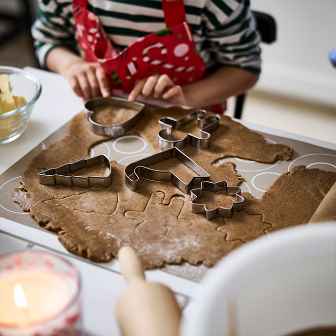 Child using metal cookie cutters with Christmas shapes (heart, reindeer, Santa hat, and star) to cut gingerbread dough on a silicone baking mat.