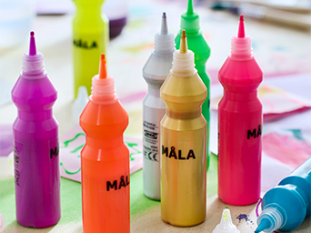 Close-up of MÅLA finger paint bottles in neon and gold colors with a dispensing tip, on a surface covered with children's drawings.