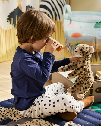 Niño sentado en el suelo, con pijama de lunares, usando binoculares de juguete mientras sostiene un peluche de leopardo. Fomenta el juego de rol y la imaginación en el espacio infantil.