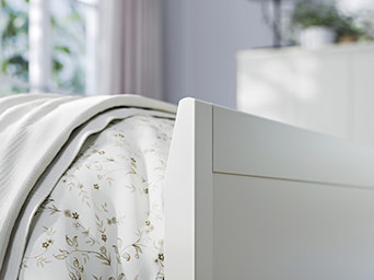 Detail of the side of a white bed with a tall headboard. The bedding is cream-patterned and covered with a blanket.
