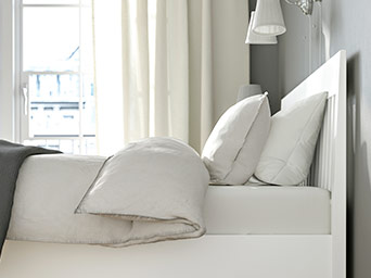 Close-up of a white bed with a slatted headboard, covered with a light duvet and fluffy pillows. Plenty of natural light.