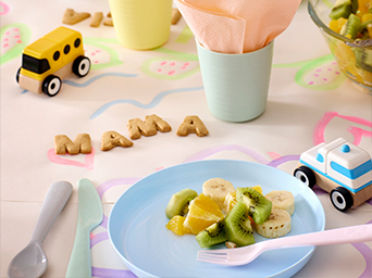 Kids' table set for snack time: Blue plate with fresh fruit (banana, kiwi, orange), colorful utensils, letter cookies ("MAMA"), and wooden toy cars. Cheerful setting for children's meals.