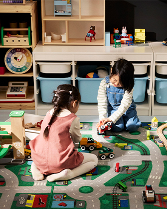 Two children playing with toy cars on a road play mat in an organized playroom with storage bins and shelves