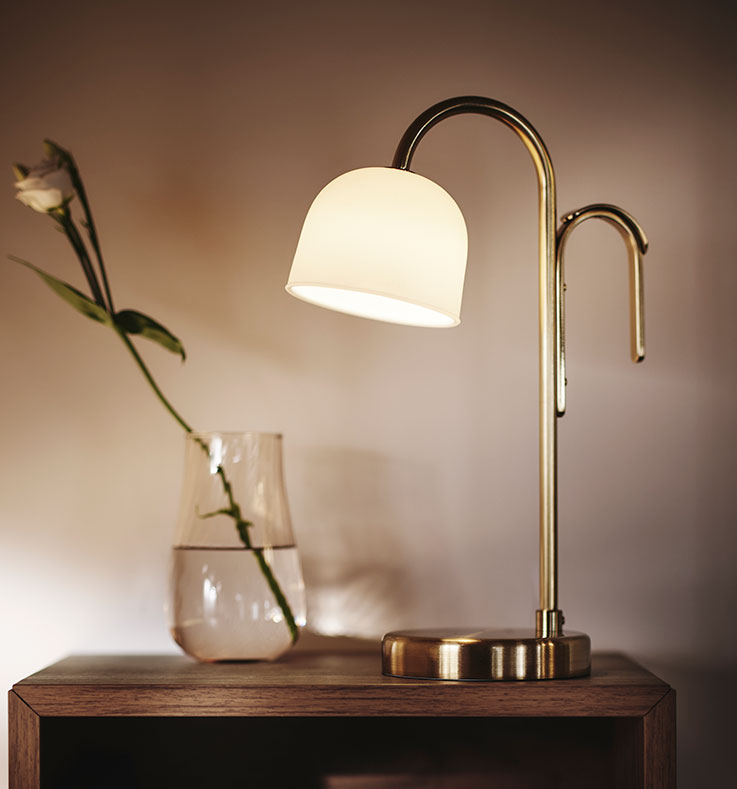 Primer plano de la lámpara portátil LED TVÅMASTAD delante de un jarrón de cristal con una flor. Luz suave alrededor.