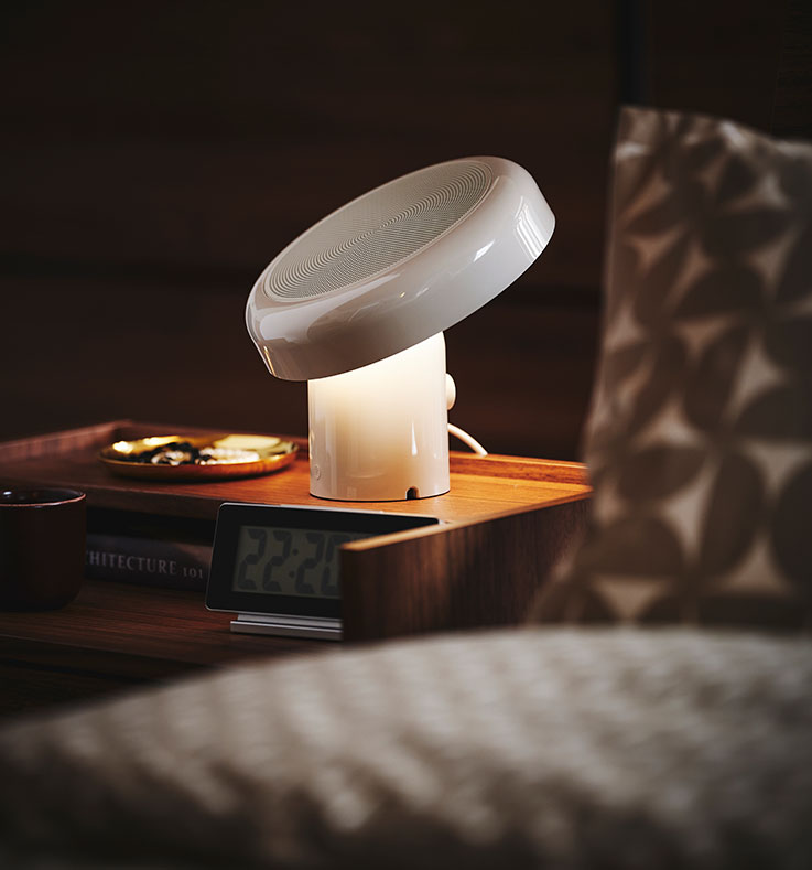 Primer plano de una mesita de noche con una lámpara con altavoz Bluetooth BLOMPRAKT, que es la única fuente de luz en la habitación, por lo demás a oscuras.