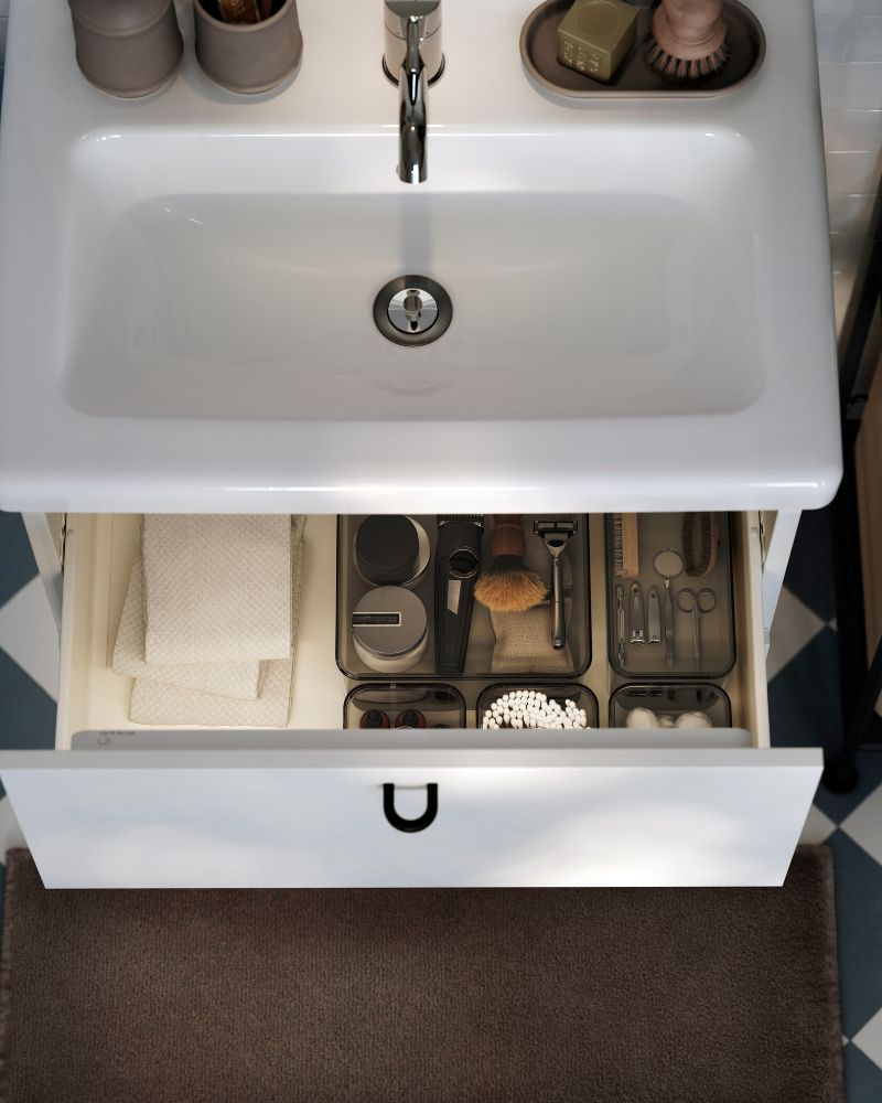Vista aérea de un mueble de lavabo ENHET/TVÄLLEN en color blanco. El cajón superior está abierto y muestra cajas con productos de afeitado.