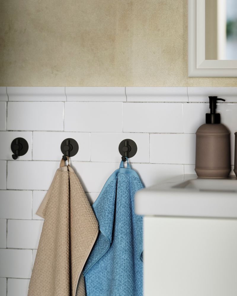 Una toalla de mano GULVIAL de color azul oscuro-gris cuelga de uno de los tres ganchos en una pared alicatada junto a un lavabo blanco en un baño.