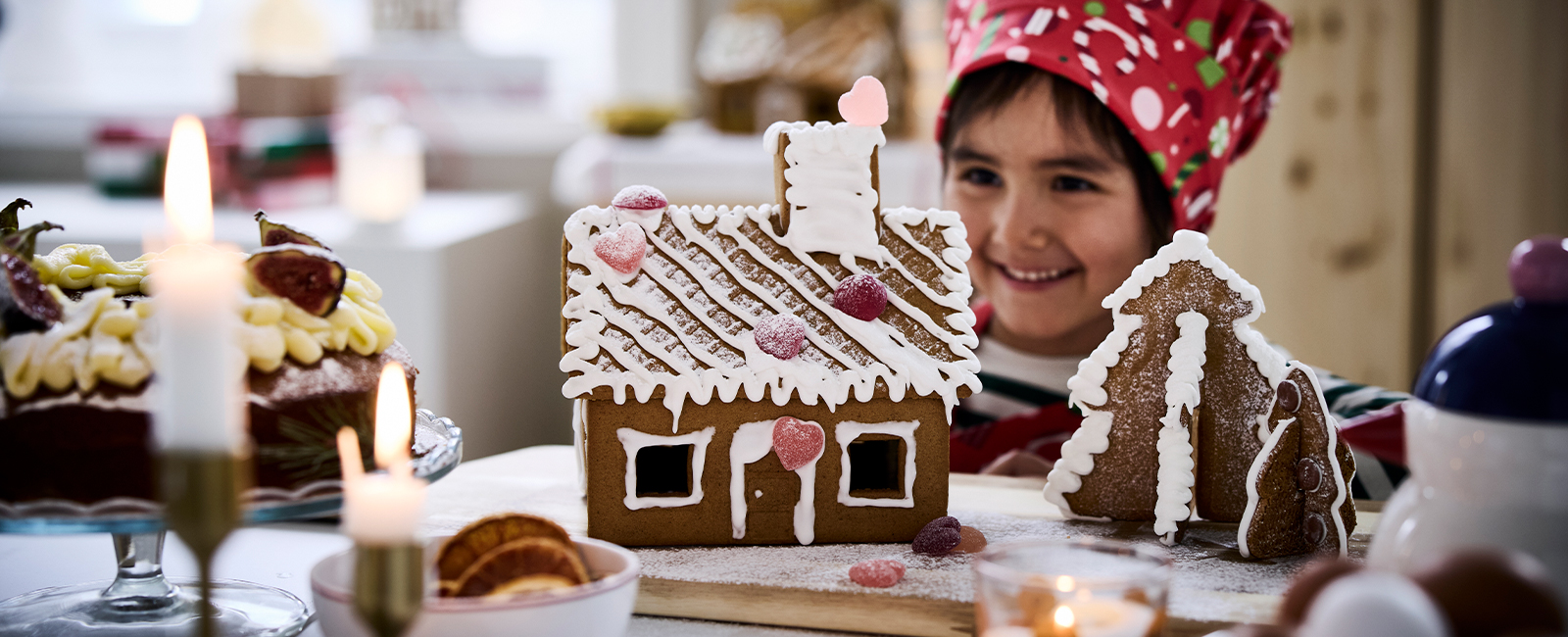 Niña decorando casa de jengibre con glaseado blanco y dulces de colores en cocina festiva con velas y postres navideños