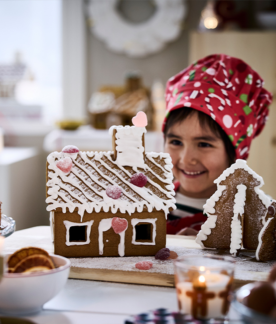 Niña decorando casa de jengibre con glaseado blanco y dulces de colores en cocina festiva con velas y postres navideños