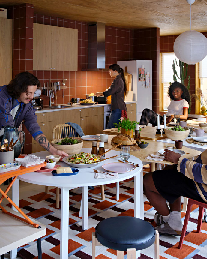 Amigos reunidos en una cocina-comedor de tonos cálidos, sirviendo pasta y ensalada en una mesa blanca para una cena social.
