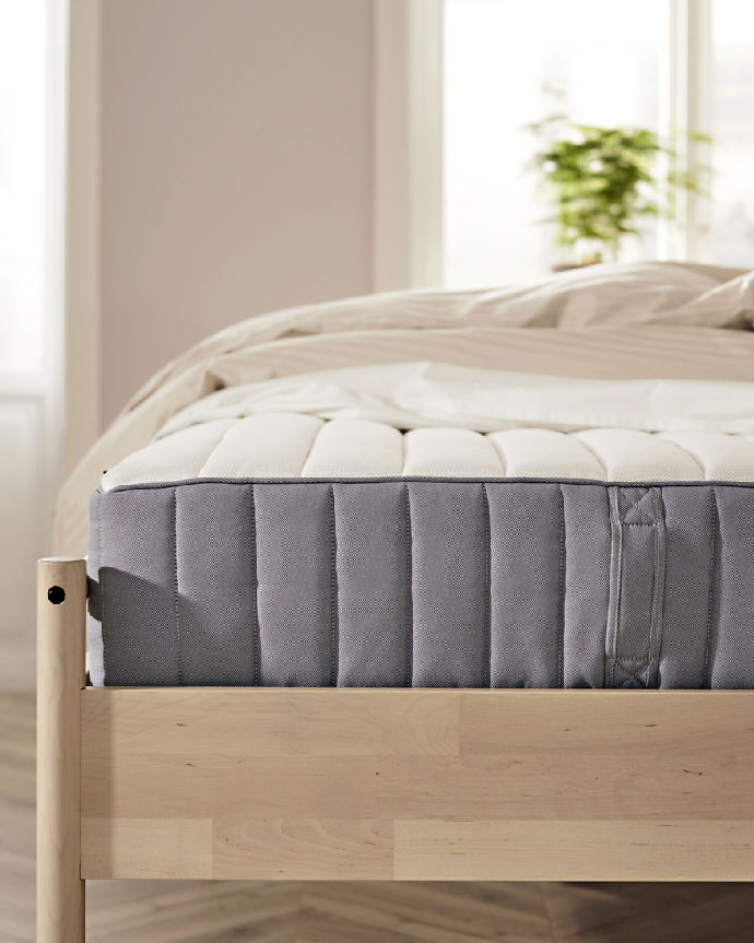 Colchón alto de color gris sobre una cama de madera clara en un dormitorio iluminado con una planta en el fondo.