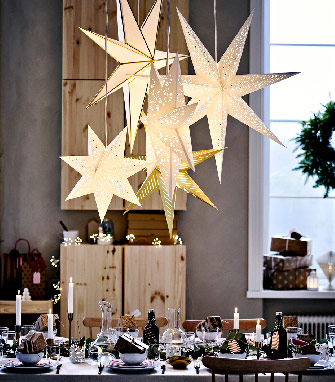 Lámparas de estrella de papel colgantes iluminan una mesa navideña puesta con platos, velas y decoraciones.