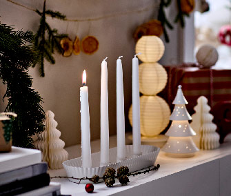 Candelabro de Adviento blanco con una vela encendida, piñas y árboles decorativos de cerámica en una repisa.