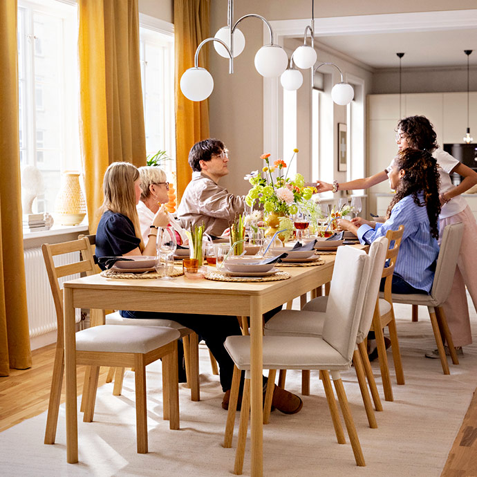 Family and friends sharing dinner at a light-wood dining table. Six people are laughing and serving food in a warm light.