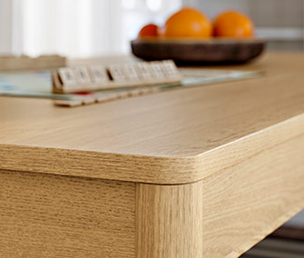 Detail of the corner of an oak wood dining table with a thick border and legs. Blurry objects are visible on the surface.