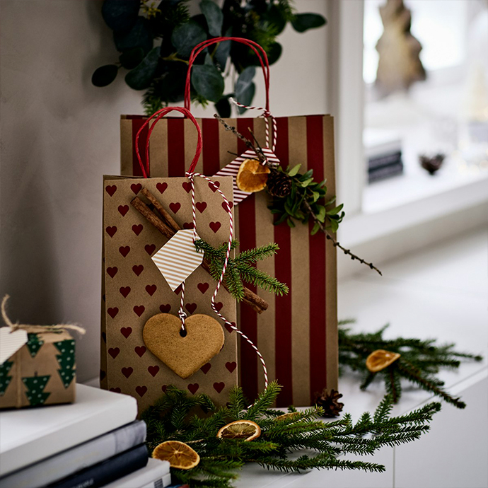 Gift bags in kraft paper with red stripes and heart patterns, decorated with cinnamon sticks, dried orange slices and evergreen sprigs. Red twisted paper handles complete the festive wrapping