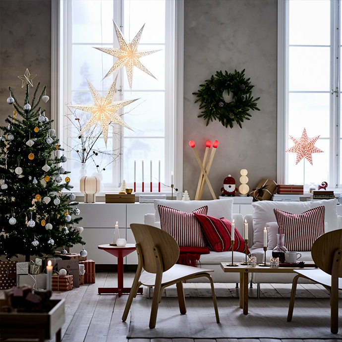 Sala de estar con árbol de Navidad, estrellas de papel en la ventana, sofá blanco con cojines a rayas rojas y mesa de centro de madera. Decoración navideña sencilla con luces y materiales naturales