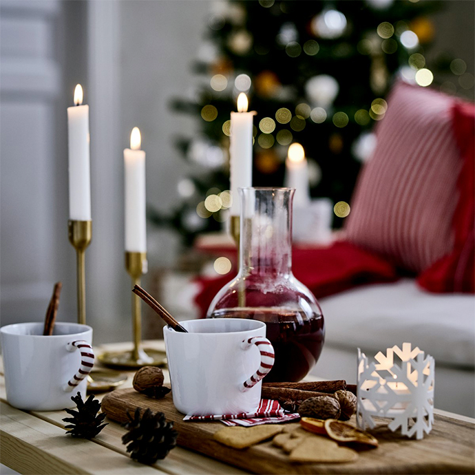 Velas blancas en portavelas de latón, bebidas calientes en tazas a rayas y decoraciones navideñas sobre superficie de madera con árbol de Navidad al fondo