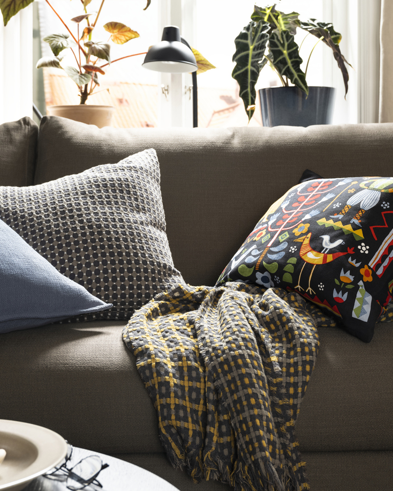 A HYLTARP sofa with a check throw and some extra cushions, including one with a multicolour TRATTKAKTUS cushion cover.