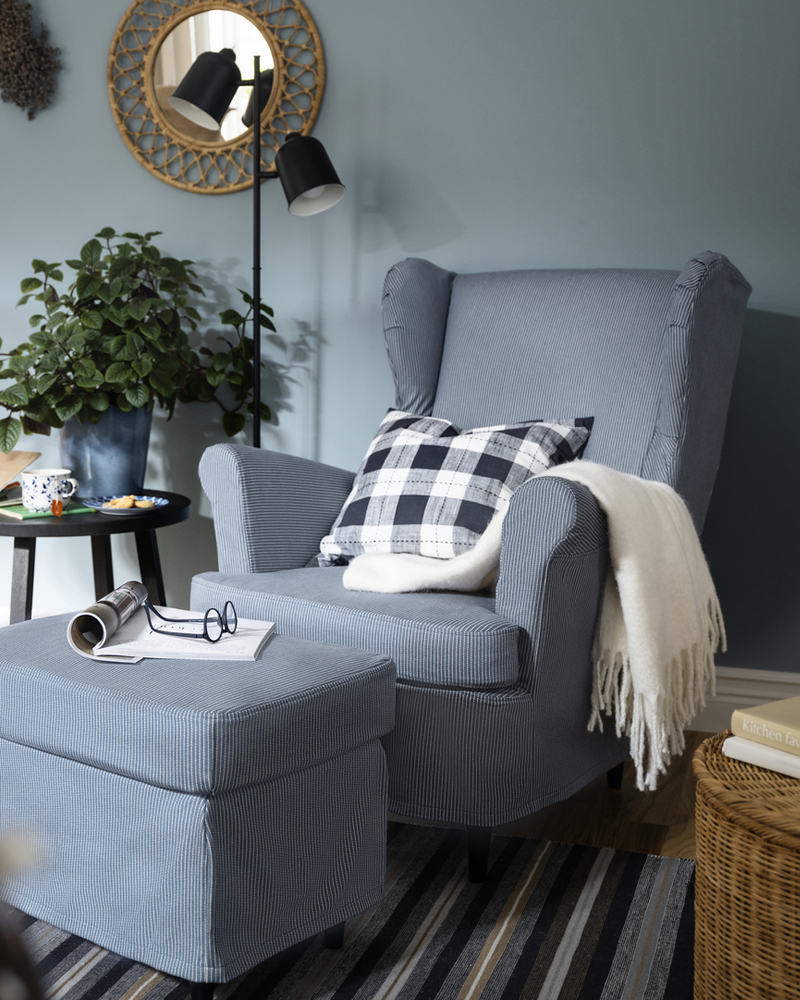 A KLÖVING oor lamp stands near a STRANDMON wing chair and footstool with a slipcover with a dark blue/beige stripe pattern.