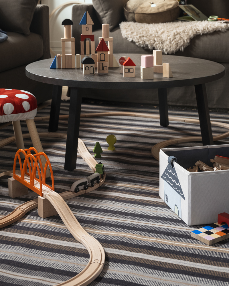 A STOCKHOLM 2025 coffee table, toys and a FLISAT children’s stool with a cover on it stand on a RÅVAROR rug in a living room.
