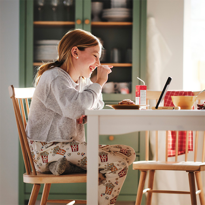 una chica joven en pijama, sentada desayunando y viendo la tablet a la vez sobre la mesa