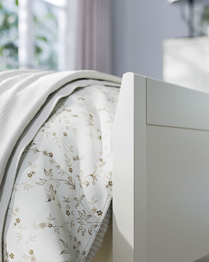 Primer plano del cabecero blanco de una cama. La ropa de cama floral y clara se ve suavemente doblada cerca de una ventana.