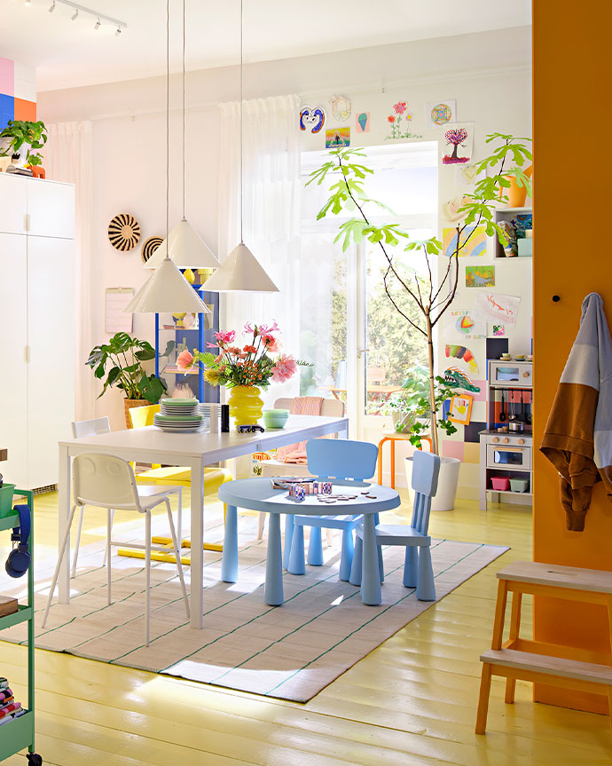 Comedor luminoso y colorido con mesa blanca y área de juego para niños. Piso amarillo, grandes ventanas y dibujos infantiles en la pared.