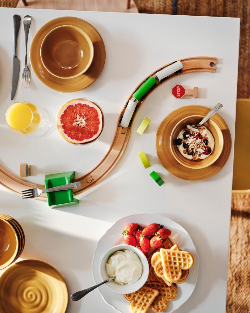 Top shot of a breakfast table set with a plate of waes and several bowls. A toy train track snakes between them.