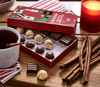 Caja de bombones de chocolate VARIADO VINTERSAGA abierta junto a una taza de té y ramas de canela sobre una tabla.