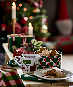 Taza de cerámica con patrón navideño, bebida caliente y galletas de jengibre con forma de corazón sobre tabla de madera.