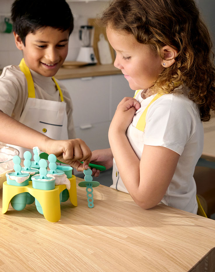 Un niño y una niña de pie junto a la encimera de la cocina preparando su propio helado con la ayuda de los moldes para polos UPPFYLLD.