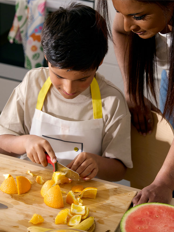 Un niño pequeño corta cuidadosamente una naranja con la ayuda del cuchillo SMÅBIT, mientras su madre lo observa desde un lado.