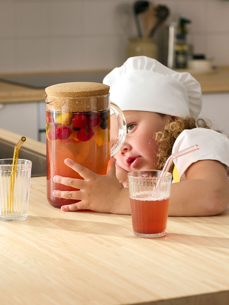 Una niña pequeña vestida con un delantal y un gorro de chef, mirando con nostalgia una jarra de agua y muchas bayas y frutas.