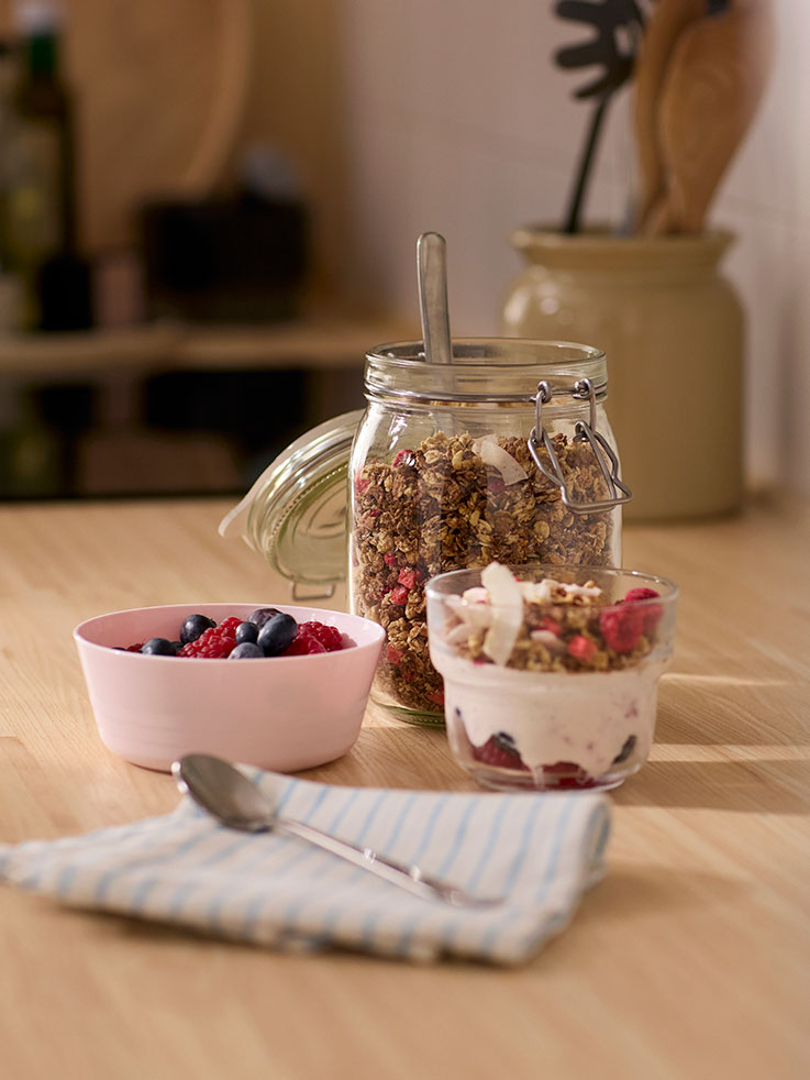 Una niña pequeña vestida con un delantal y un gorro de chef, mirando con nostalgia una jarra de agua y un montón de bayas y frutas.