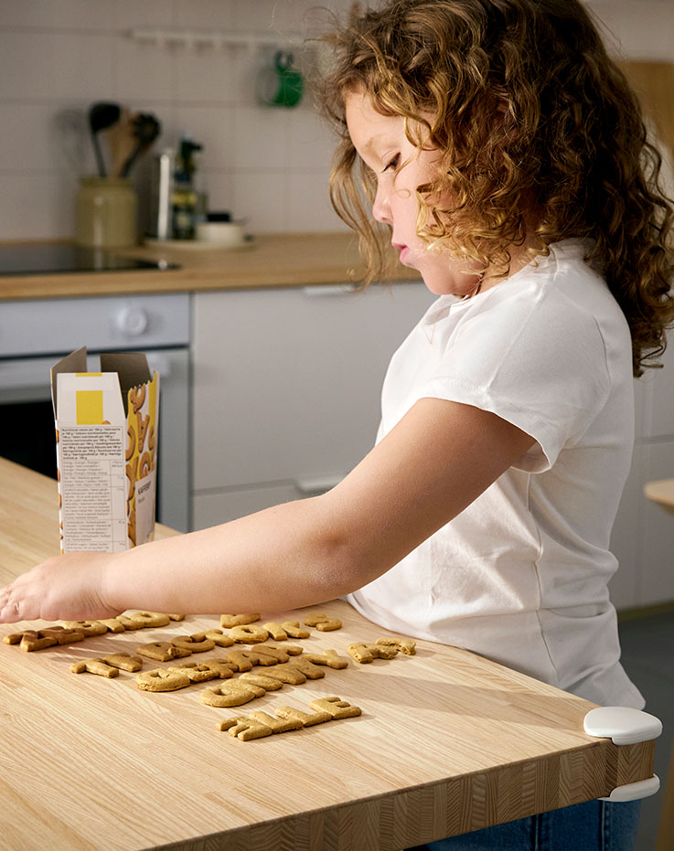 Una niña pequeña utiliza galletas KAFFEREP para escribir diferentes nombres en la encimera de la cocina. Protector de esquinas UNDVIKA colocado.