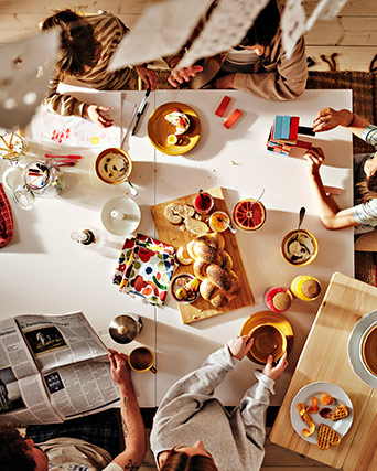 Top view of shared breakfast with bread, fruit, and newspaper. Several people around the table.