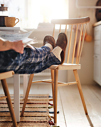 Close-up of feet in slippers and socks, resting on a light wood dining chair. The person is wearing blue plaid pajamas.